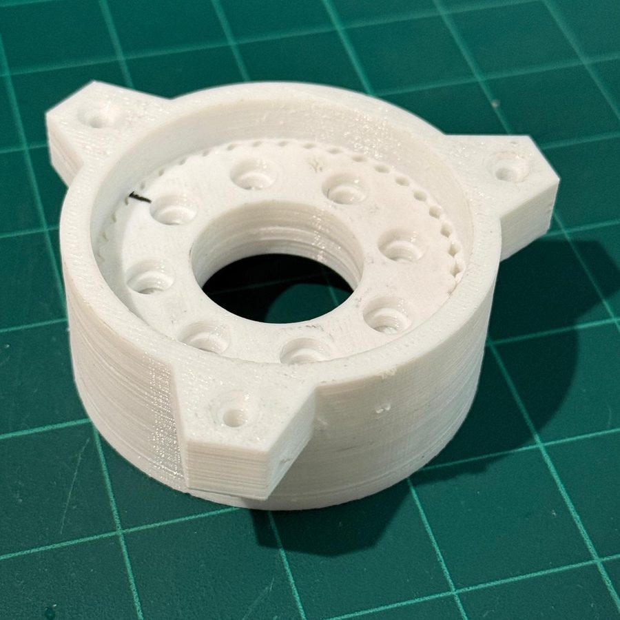 White circular housing with three mounting lugs, a ring of counterbored holes around a finely ridged inner ring, and a large central bore